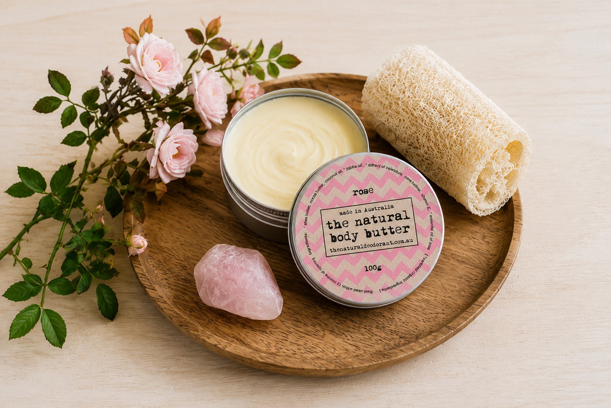 A round tin of rose body butter with a pink label sits on a wooden tray beside an open jar showing pale yellow cream, a rolled natural loofah, and a pink crystal on a light wood surface with pink roses and green leaves.