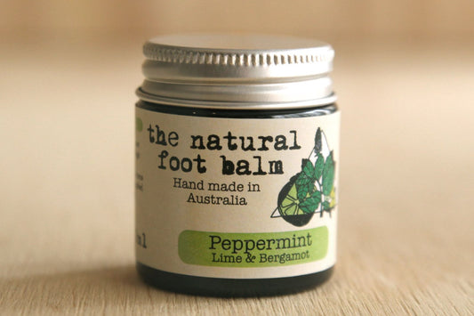 Jar of 'the natural foot balm' with peppermint, lime, and bergamot on a wooden surface.