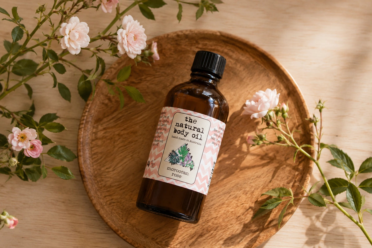 A bottle of 'the natural body oil' botanical body oil with a black cap and a pink-and-white patterned label, resting on a round wooden tray surrounded by pink roses and green leaves on a light wood table.