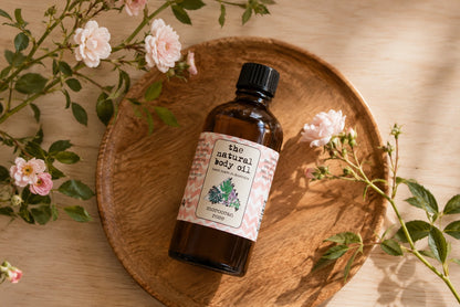 A bottle of 'the natural body oil' botanical body oil with a black cap and a pink-and-white patterned label, resting on a round wooden tray surrounded by pink roses and green leaves on a light wood table.